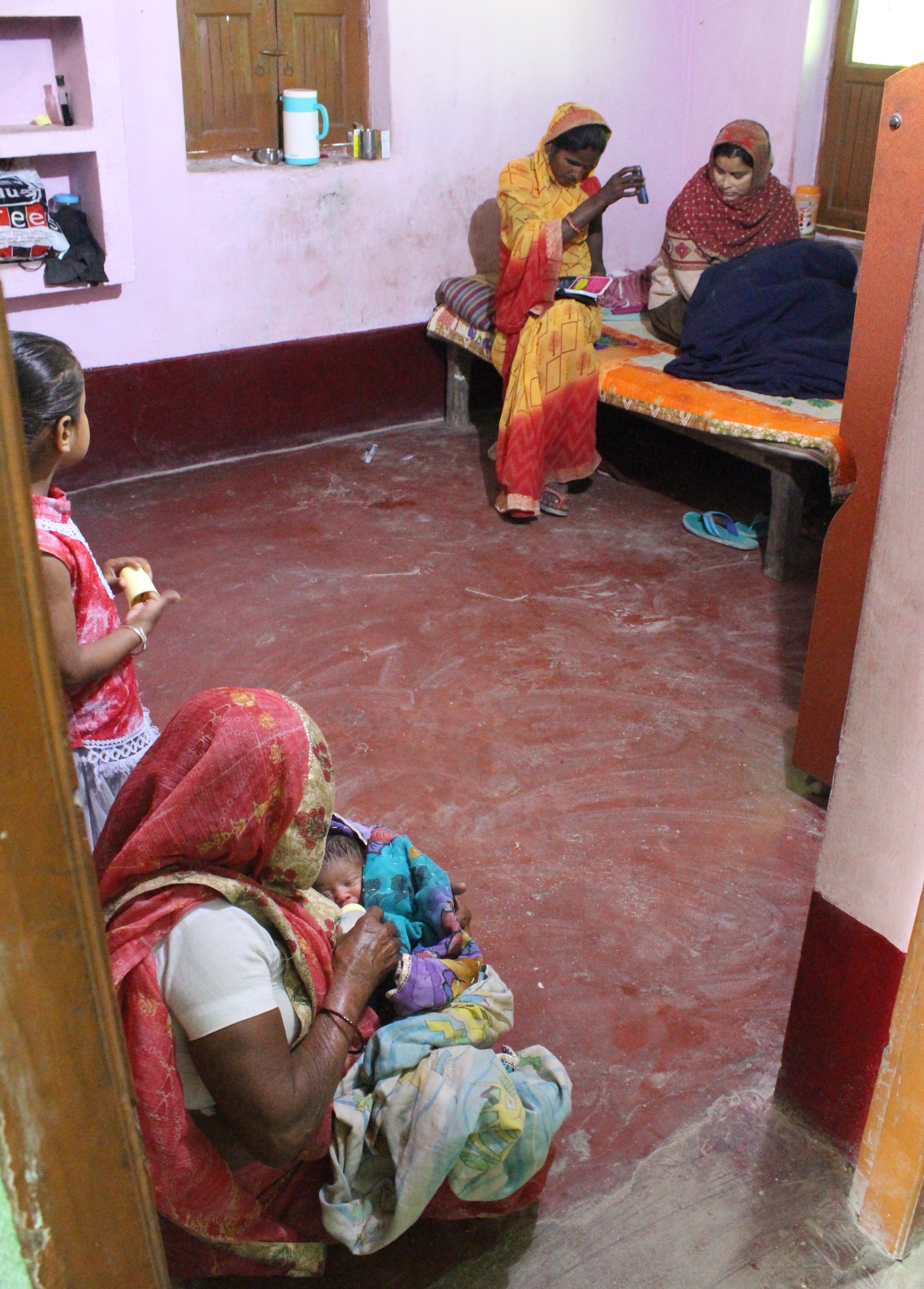 An ASHA plays a recording from her mobile phone about the importance of breast-feeding to a mother, while the mother's new baby is being bottle-fed in the foreground, potentially showing a contrast between an emphasis on service extension and cultural facilitation.